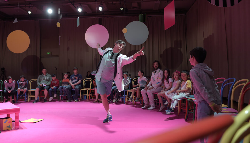 An Invisible Orchestra production photo. A performer in a white suit jacket stands on pink carpet, gesturing with a flourish. There is a circle of chairs around him, with families sitting and standing. Large colourful shapes are suspended from the ceiling.