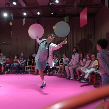 An Invisible Orchestra production photo. A performer in a white suit jacket stands on pink carpet, gesturing with a flourish. There is a circle of chairs around him, with families sitting and standing. Large colourful shapes are suspended from the ceiling.