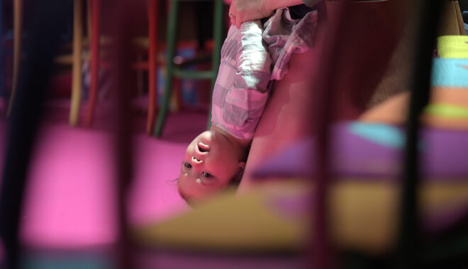 An Invisible Orchestra production photo. A child is suspended upside down by their adult, visible between the backs of colourful chairs.