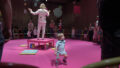 An Invisible Orchestra production photo. A small child stands in the centre of an expanse of pink carpet. On the outskirts is a circle of chairs, with families sitting or standing. A performer in a white suit stands on a small pink platform, his arms raised and his back to the camera. Large colourful shapes are suspended from the ceiling.