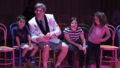 An indoor Invisible Orchestra production photo. A performer in a white suit jacket sits on a chair, tilting his head to see something out of frame. Three children sit on chairs around him. The scene is illuminated with pinkish theatre lighting.