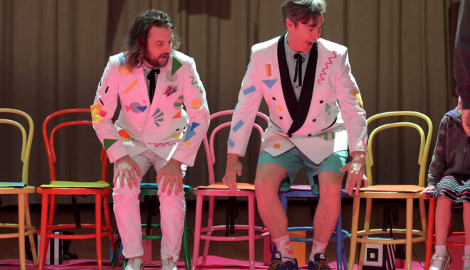 An indoor Invisible Orchestra production photo. Two performers in white patterned suit jackets are about to sit on colourful chairs. They are both looking with interest at an audience member out of frame.