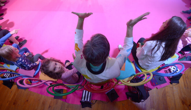 An indoor Invisible Orchestra production photo. An overhead shot of a performer in a white jacket, sitting on a colourful chair, gesturing wildly with his hands. Children and adults sit either side of him, and one child is looking up into the camera.