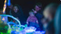 A Light Pickers production photo. Two adults sit with their babies on their laps behind a pile of abstract glowing objects. The adults are gesturing to something out of frame, showing their babies.