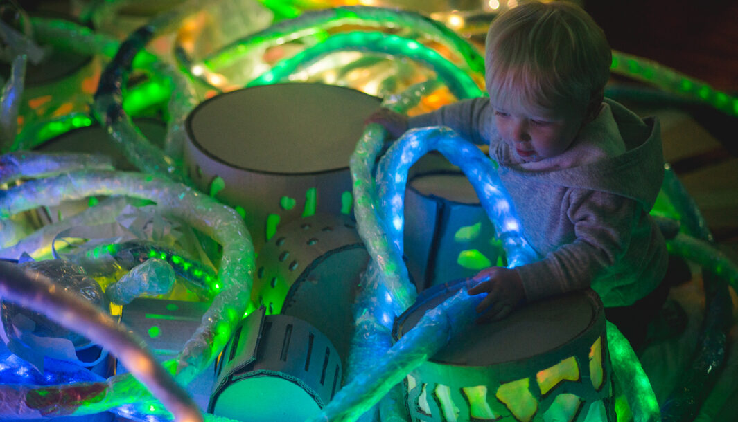 A Light Pickers production photo. A small child is surrounded by abstract glowing objects, which they are interacting with interestedly.