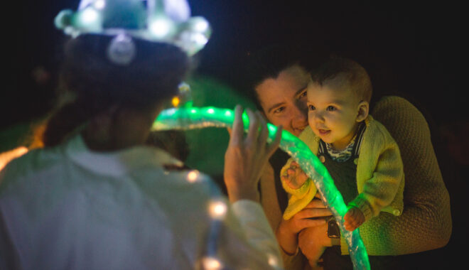 A Light Pickers production photo. An adult holds their baby, who is looking at a Polyglot artist moving an abstract glowing green object. The artist wears a white boiler suit and a glowing helmet and has their back to the camera. They are in a darkened space.