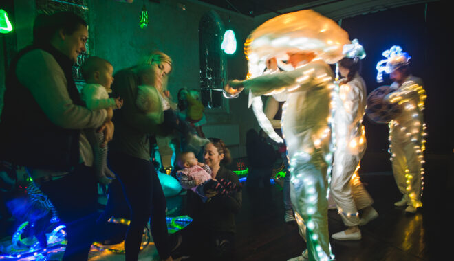 A Light Pickers production photo. Adults holding their babies stand in a darkened space, interacting with three Polyglot artists in white boiler suits, draped in strands of light. A large, abstract glowing object is suspended from the ceiling.