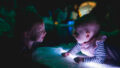 A Light Pickers production photo. A small child and a baby lie on an abstract glowing object, smiling at each other. Other objects glow in the background. They are in a darkened space.