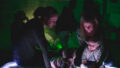 A Light Pickers production photo. Two adults and their babies play with abstract glowing objects. They are in a darkened space, and the area behind them is illuminated with green light.