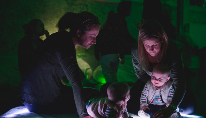 A Light Pickers production photo. Two adults and their babies play with abstract glowing objects. They are in a darkened space, and the area behind them is illuminated with green light.