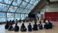 A professional development photo. A group of artists sit in a circle. They are in a large space, with a sloped glass ceiling and wooden floors. Photo courtesy of Esplanade - Theatres on the Bay, Singapore