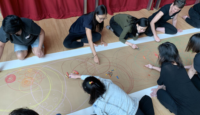 A professional development photo. Artists sit around a long piece of brown paper on a wooden floor, drawing.