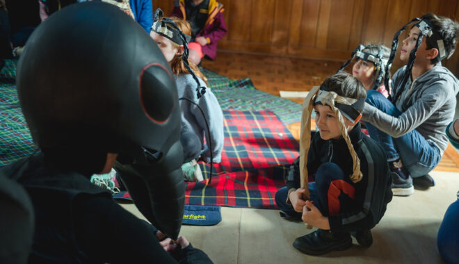 An indoor Ants production photo. A Polyglot artist in an intricate Ant costume crouches on the floor, engaging with a child wearing handmade paper antennae. Other children and families are visible in the background.