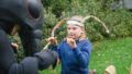 Ants production photo: Theresa Harrison. A Polyglot artist in an intricate black Ant costume engages with a young person wearing handmade paper antennae. They are outdoors on a green lawn, next to large shrubs.
