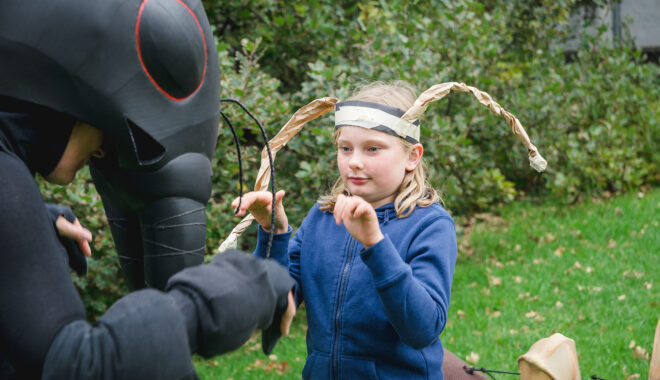 Ants production photo: Theresa Harrison. A Polyglot artist in an intricate black Ant costume engages with a young person wearing handmade paper antennae. They are outdoors on a green lawn, next to large shrubs.