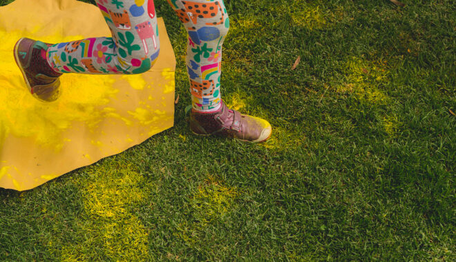 A Bees production photo. An overhead shot of a child's legs and feet running through yellow 'pollen' on green grass. The child wears colourful patterned leggings.