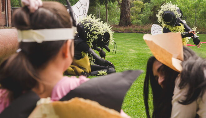A Bees production photo. A child and an adult in handmade paper bee costumes watch Polyglot artists who wear intricate black and yellow Bee costumes. They are on a green lawn.
