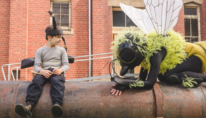 A Bees production photo. A Polyglot artist in an intricate Bee costume is perched on a large metal pipe. A child wearing handmade paper antennae and wings sits on the pipe looking at them. There is a red brick building in the background.