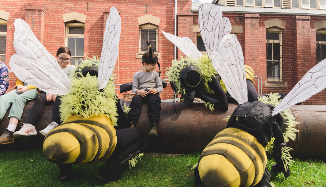 A Bees production photo. Polyglot artists in intricate Bee costumes are perched on and around a large metal pipe on green lawn. Children wearing handmade paper antennae and wings sit on the pipe amongst them. There is a heritage red brick building in the background.