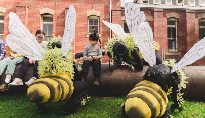 A Bees production photo. Polyglot artists in intricate Bee costumes are perched on and around a large metal pipe on green lawn. Children wearing handmade paper antennae and wings sit on the pipe amongst them. There is a heritage red brick building in the background.