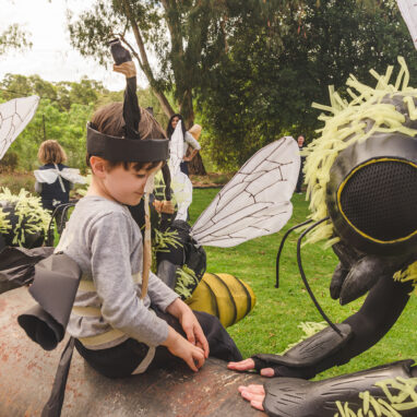 A Bees production photo. Polyglot artists in intricate Bee costumes are perched on and around a large metal pipe. A child wearing handmade paper antennae and wings sits on the pipe looking at them. There is green lawn and trees in the background.
