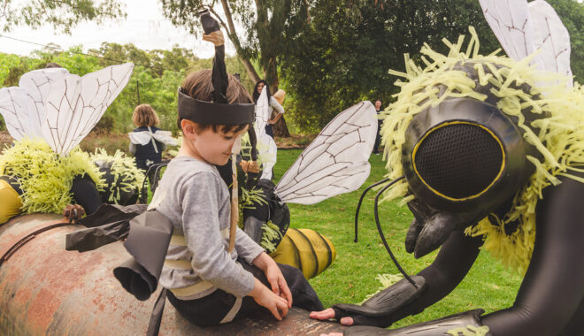 A Bees production photo. Polyglot artists in intricate Bee costumes are perched on and around a large metal pipe. A child wearing handmade paper antennae and wings sits on the pipe looking at them. There is green lawn and trees in the background.