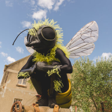 A Bees production photo. A Polyglot artist, wearing an intricate black and yellow Bee costume, perches on a metal object. A green tree and heritage buildings are in the background. Blue sky with sparse white clouds stretches behind them.