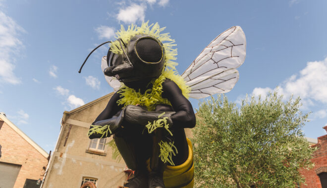 A Bees production photo. A Polyglot artist, wearing an intricate black and yellow Bee costume, perches on a metal object. A green tree and heritage buildings are in the background. Blue sky with sparse white clouds stretches behind them.