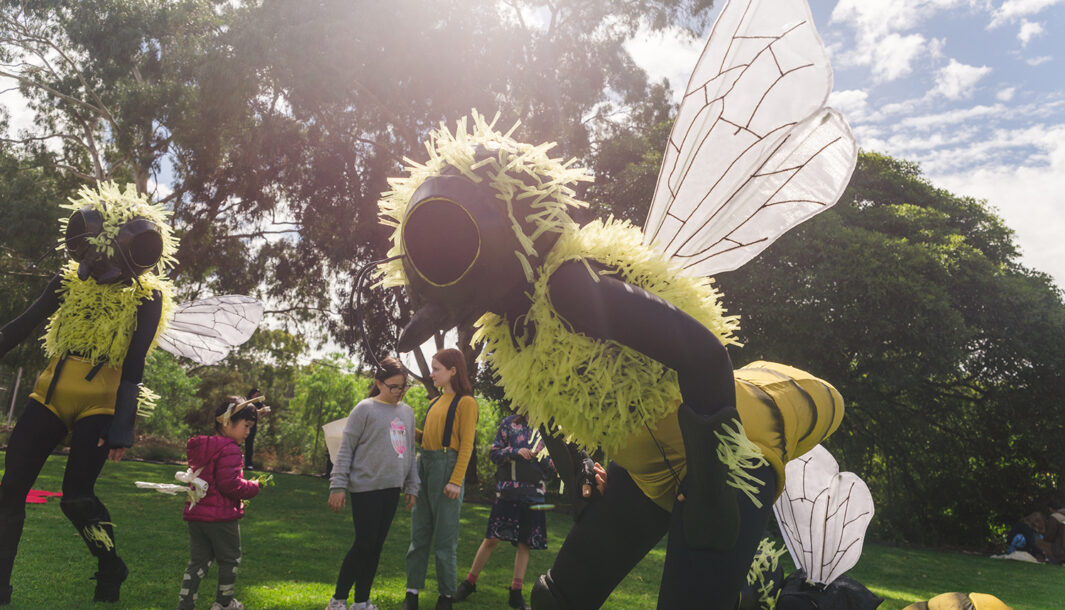 A Bees production photo. A Polyglot artist in an intricate Bee costume stands in a stylised position. In the background are children and other artists in Bee costumes. They are on a green lawn, with trees and blue sky in the background. Photo: Theresa Harrison