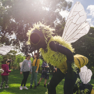 A Bees production photo. A Polyglot artist in an intricate Bee costume stands in a stylised position. In the background are children and other artists in Bee costumes. They are on a green lawn, with trees and blue sky in the background.