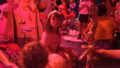 A Paper Planet production photo. A child sits among tall cardboard trees, pulling a cheeky face at another child. The scene is illuminated with pinkish-red theatre lighting.