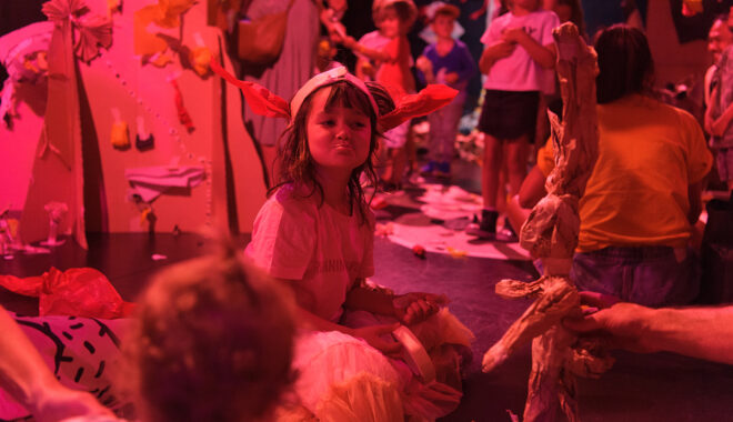 A Paper Planet production photo. A child sits among tall cardboard trees, pulling a cheeky face at another child. The scene is illuminated with pinkish-red theatre lighting.