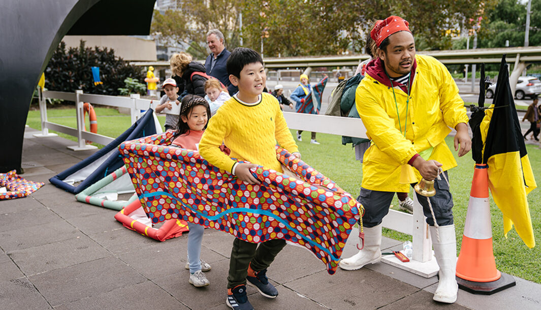 An outdoor Boats production photo. A performer wearing gumboots and a raincoat rings a bell. To their right, two children run forwards in a bottomless boat-shaped vessel on a tiled concrete pavement.