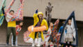 An outdoor Boats production photo. A Polyglot artist in a yellow raincoat, rain hat, white gumboots and orange flotation ring is shouting and gesturing excitedly from a set of stairs next to a statue. They are surrounded by excited, shouting children holding colourful vessels.