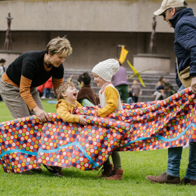 An outdoor Boats production photo. Children and their adults play with colourful patterned vessels. They are on green grass with a building in the background.