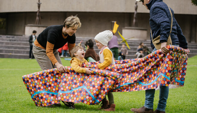 An outdoor Boats production photo. Children and their adults play with colourful patterned vessels. They are on green grass with a building in the background.