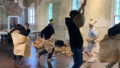 A professional development photo. Polyglot artists, wrapped in brown paper, or wearing handmade brown paper costumes, move and dance in a room with wooden floors. Photographer: Lexie Wood