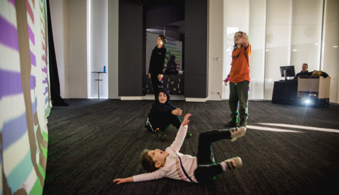 A Sound Shadows production photo. Three children and an adult engage with their shadows on a large screen with brightly coloured projections on it. One of the children is lying on the floor, another is seated on the floor, while the third child and the adult are standing. Photographer: Theresa Harrison