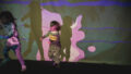 A Sound Shadows production photo. Two children play in front of a large screen with projections and shadows on it. Photographer: Theresa Harrison