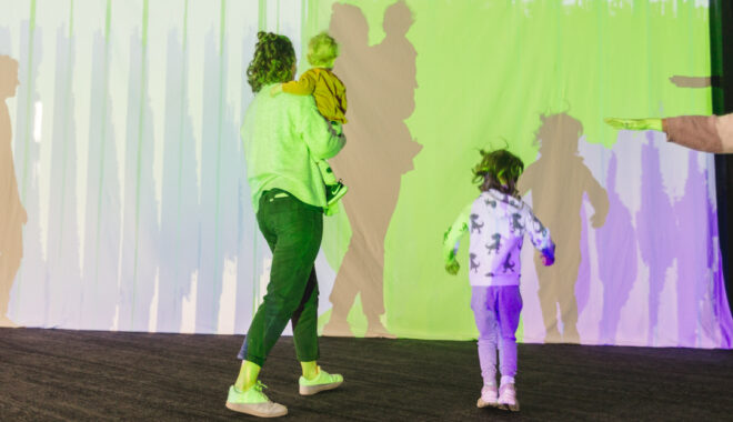 A Sound Shadows production photo. A parent and two children stand in front of colourful projections on a screen. The mother is holding one of her children, and the other child stands beside her Photo: Theresa Harrison.