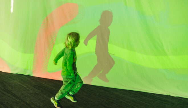 A Sound Shadows production photo. A small child runs in front of colourful projections on a screen. Photo: Theresa Harrison.