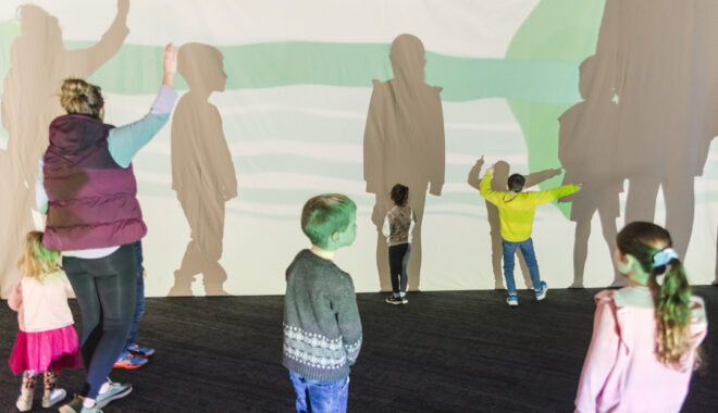 A Sound Shadows production photo. A group of children pose to make shadows in front of colourful projections on a screen. Photo: Theresa Harrison.