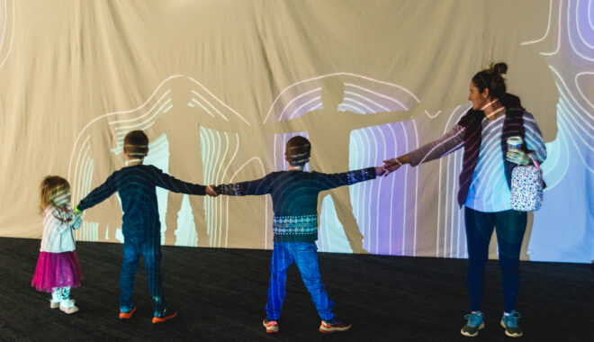 A Sound Shadows production photo. Three children and an adult hold hands to engage with their shadows which fall on a screen onto which wavy lines and colours are projected. Photographer: Theresa Harrison