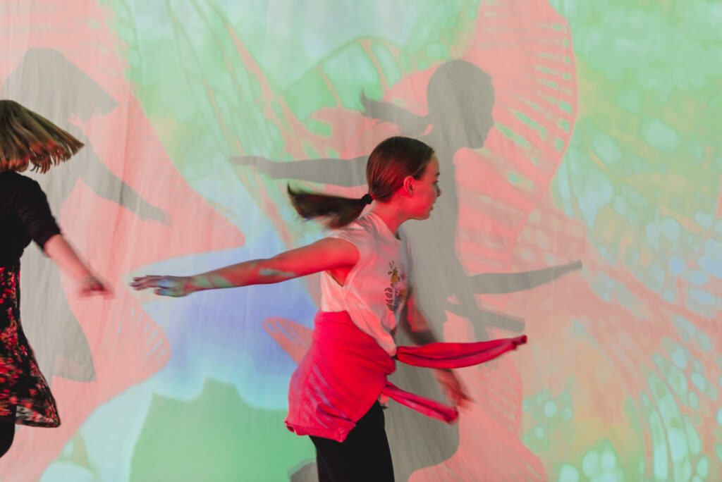 A Sound Shadows production photo. A child with a ponytail plays in front of a screen with red, green and blue coloured images on it. The child has one arm outstretched behind her and the other one in front of her. Photographer: Theresa Harrison