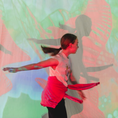 A Sound Shadows production photo. A child with a ponytail plays in front of a screen with red, green and blue coloured images on it. The child has one arm outstretched behind her and the other one in front of her. Photographer: Theresa Harrison