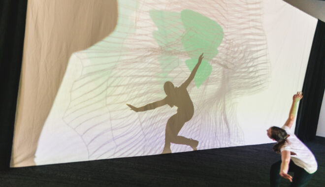 A Sound Shadows production photo. A child poses with their legs knelt and their arms outstretched in front of a colourful screen to make shadows. Photo: Theresa Harrison.
