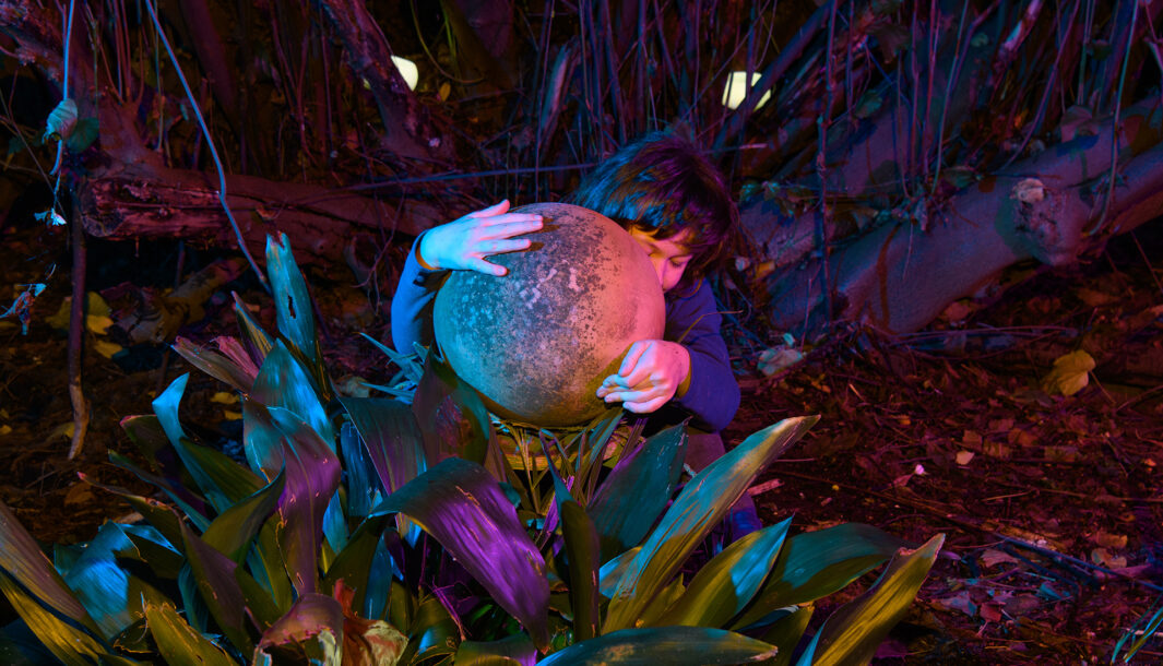 A When the World Turns photo. A child sits amongst plants and trees, clasping a large spherical object that partially covers their face. It is night and the image is dark, with deep purple tones. Photographer: Sarah Walker