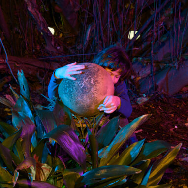 A When the World Turns photo. A child sits amongst plants and trees, clasping a large spherical object that partially covers their face. It is night and the image is dark, with deep purple tones. Photographer: Sarah Walker