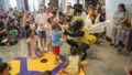 A Bees production photo. A child stands on a yellow mat with their hands over their head. A performer in a bee costume kneels towards them with their hands stretched towards the child. Photo: Alvaro Keding.