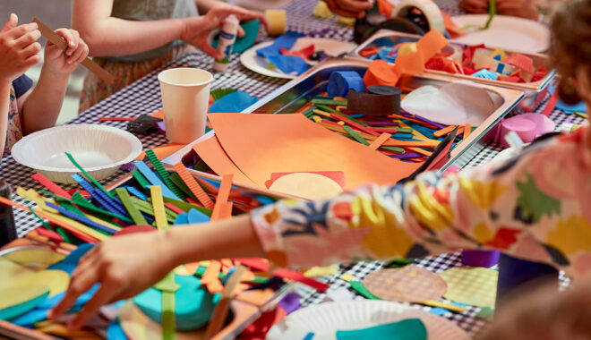 A Feast Production photo. Children surround a table, with metal tubs full of coloured pencils, and coloured paper. A child's arm reaches out to a tub to grab something. Photo: Casey Horsfield.